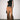 A model stands against a plain light gray wall wearing a black sleeveless mock-neck top and wide-leg brown satin pants with lace trim at the hems, styled with black pointed shoes and holding a small dark clutch.