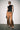 A model stands against a plain light gray wall wearing a black sleeveless mock-neck top and wide-leg brown satin pants with lace trim at the hems, styled with black pointed shoes and holding a small dark clutch.
